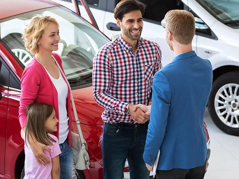 A family shakes hands with a man and woman in a car showroom, surrounded by various vehicles on display.